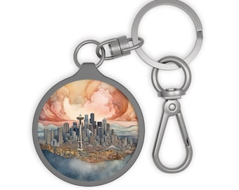 Puede incluir: Un llavero plateado con un dije redondo que presenta una ilustración de acuarela del horizonte de Seattle con la Space Needle. El dije tiene un borde plateado.