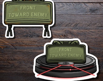 Könnte beinhalten: Zwei Aufkleber. Einer zeigt ein grünes rechteckiges Objekt mit dem Text "FRONT TOWARD ENEMY" und Beinen. Der andere zeigt dasselbe Objekt auf einer schwarzen kreisförmigen Basis mit roten Laserlinien, auf dunklem Holzmaserungshintergrund.