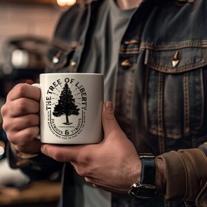 May include: White ceramic mug with a black graphic of a tree and the text "THE TREE OF LIBERTY" and "PATRIOTS & TYRANTS" with a quote by Thomas Jefferson. The mug is held by a person wearing a denim jacket.