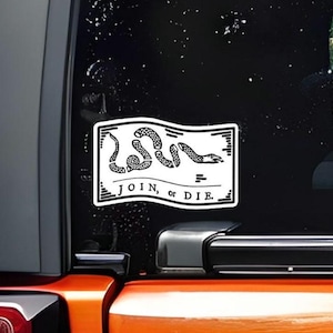 May include: A white rectangular sticker with a black outline, featuring a coiled snake and the words "JOIN, or DIE." The sticker is affixed to a vehicle's window, with the background showing a glimpse of a green lawn.