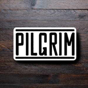 May include: A rectangular sticker with the word "PILGRIM" in bold, black letters. The text is centered on a white background, framed by a thick black border. The sticker is set against a dark wood grain background.