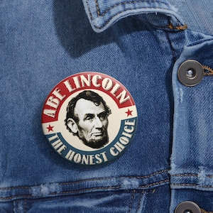 Puede incluir: Un botón redondo con un retrato de Abraham Lincoln, el texto "ABE LINCOLN" en rojo y "THE HONEST CHOICE" en azul. El botón está sobre una chaqueta vaquera.