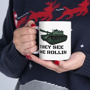 May include: White ceramic mug featuring a green tank illustration with the text "THEY SEE ME ROLLIN" in black. The mug has a curved handle and is held by a person.