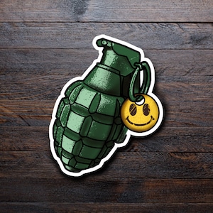 May include: A green grenade sticker with a white border and a yellow smiley face charm. The grenade has a detailed design with a ring and pin. The smiley face charm has a black smiling face.