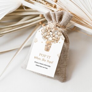 Könnte beinhalten: Ein kleiner Jutesack, der mit Bindfaden zusammengebunden ist und ein Etikett mit einem Heißluftballon-Design trägt. Auf dem Etikett steht "POP IT When She Pops!" mit einem Teddybär im Korb. Der Text darunter lautet "Stoßen Sie an, wenn Baby Robertson kommt."