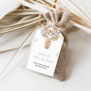 May include: A small hessian bag tied with twine, with a tag showing a hot air balloon design. The balloon is filled with blue and gold orbs, with a teddy bear in the basket. The tag reads "POP IT When She Pops!" and includes a personalized message.