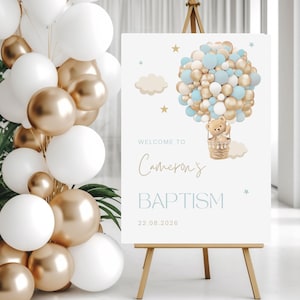 以下が含まれることがあります： キャメロンの洗礼式用のウェルカムサイン。熱気球のデザインとバスケットに入ったテディベアが特徴です。イーゼルに立てかけられ、背景には風船のガーランドがあります。日付22.08.2026が含まれています。