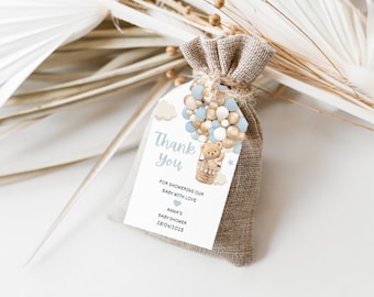 May include: A white thank you tag with a blue and gold hot air balloon illustration featuring a teddy bear. The tag is attached to a small burlap bag. The text on the tag reads "Thank you for showering our baby with love Anna's Baby Shower 28/04/2023".