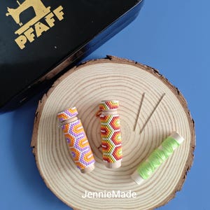 Puede incluir: Dos estuches de agujas con cuentas con patrones geométricos coloridos, una bobina de hilo verde y dos agujas descansan sobre una rodaja de madera. Una caja negra de máquina de coser con la palabra "PFAFF" está en el fondo. El texto "JennieMade" está en la parte inferior.