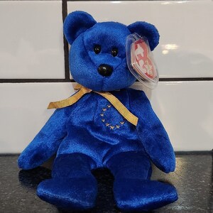 May include: A plush blue teddy bear with a gold ribbon tied around its neck. The bear has a black nose and eyes, and a tag is attached to its ear. A gold emblem of stars is on the bear's chest.