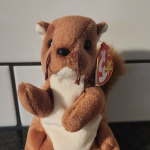 Op de afbeelding: Een bruin pluche eekhoornspeelgoed met een witte buik en een pluizige staart. De eekhoorn heeft zwarte ogen en een rood label aan zijn oor. Het speelgoed staat op een donkere ondergrond.