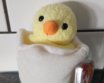 Peut inclure: Un jouet en peluche représentant un poussin jaune avec des yeux noirs et un bec orange, sortant d'une coquille d'œuf blanche. Le poussin est fait d'une matière douce et texturée. Une étiquette rouge en forme de cœur est attachée.