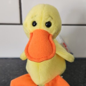 May include: A plush yellow duck toy with an orange beak and feet. The duck has black eyes and small wings. The toy is made of soft material and is designed to stand upright. A small tag is attached to the duck.