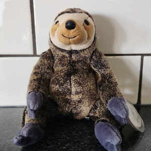 May include: A soft sloth toy featuring a brown and tan patterned body, a cream-coloured face, and dark blue feet and hands. The sloth has a smiling expression and a black nose. The toy is sitting on a dark surface.
