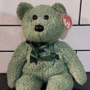 May include: A green plush teddy bear with a dark green bow and a shamrock accent. The bear has black eyes and a black nose. A red heart-shaped tag is attached to the ear.