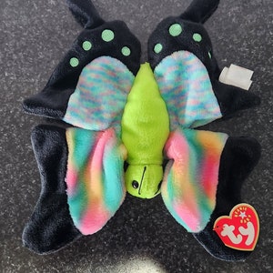 May include: A plush butterfly toy with black wings featuring green polka dots and rainbow-coloured accents. The body is lime green. A red TY Beanie Baby tag is attached.