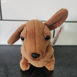 May include: A brown plush dachshund dog toy with floppy ears, black eyes, and a black nose. The stuffed animal is sitting upright and has a tag attached to its ear. The toy is on a dark surface.