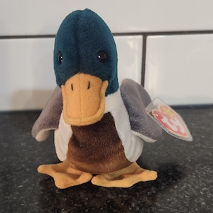 May include: A plush duck toy with a teal head, brown body, and gray wings. The duck has an orange beak and small black eyes. A small tag is attached to the duck's wing.