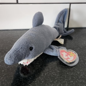 May include: A grey plush shark toy with a white belly and a red and white mouth. The shark has a fin on its back and a tag attached to its side. The tag is heart-shaped with the words "TY" and "Beanie Baby" on it.