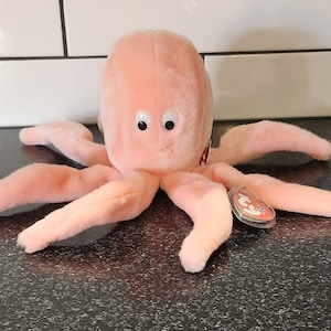 May include: A plush, peach-colored octopus toy with eight long tentacles and large, round eyes. The stuffed animal has a tag attached to one of its tentacles. The toy is sitting on a dark surface.