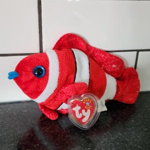 May include: A plush clownfish toy in red, white, and black. The toy has large blue eyes and a blue nose. A heart-shaped tag is attached to the toy. The toy is on a dark surface.