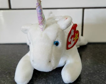 Könnte beinhalten: Ein weißes Plüsch-Einhorn-Spielzeug mit einem schimmernden, irisierenden Horn und blauen Augen. Das Einhorn hat ein rotes Etikett an seinem Ohr. Das Spielzeug liegt auf einer dunklen Oberfläche.