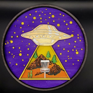 May include: A round wooden wall art piece featuring a layered design. The artwork depicts a gold UFO above a mountain scene with a disc golf basket, two aliens, and a starry purple sky. The piece is framed in a dark wood.