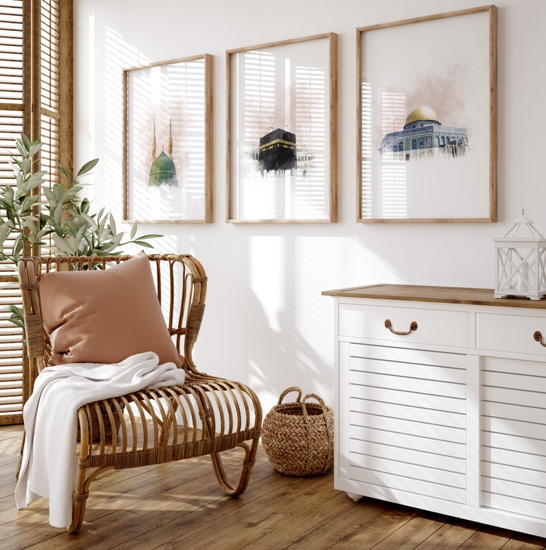 May include: A wicker chair with a brown throw blanket and a tan pillow sits in front of a white wall with three framed watercolor paintings of mosques. A white cabinet with two drawers and a wooden top is to the right of the chair.