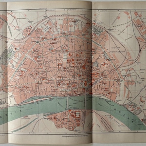 May include: Vintage map of Cologne, Germany, featuring a detailed city plan with streets, buildings, and the Rhine River. The map is printed in shades of pink, green, and brown, with text in German. The map is open, showing the city's layout.