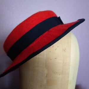 Puede incluir: Un sombrero de fieltro rojo con una banda negra alrededor de la corona. El sombrero está sentado en una cabeza de maniquí de madera.