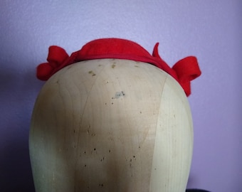 May include: A red felt hat with a large bow on the back. The hat is on a wooden hat form.