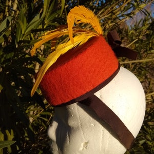 Peut inclure: Un chapeau en feutre rouge avec une plume jaune et un ruban noir. Le chapeau est sur une tête de mannequin blanche.