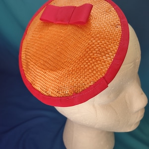 Puede incluir: Un sombrero redondo de paja naranja con un lazo de cinta rojo. El sombrero está en una cabeza de maniquí blanca.