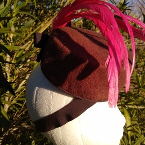 Peut inclure: Un chapeau en feutre marron avec un accent de plume rose. Le chapeau est sur une tête de mannequin blanche.