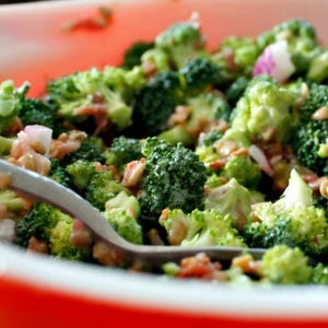 May include: A close-up of a broccoli salad in a red bowl. The salad contains bright green broccoli florets, mixed with red onion and a light dressing. A silver fork is placed in the salad. The image is well-lit and in focus.