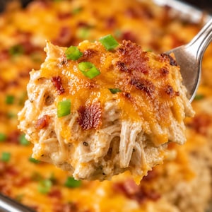 May include: A fork holds a portion of a casserole, showcasing shredded chicken, melted cheddar cheese, and crispy bacon pieces, garnished with spring onions. The dish is a warm, inviting meal.
