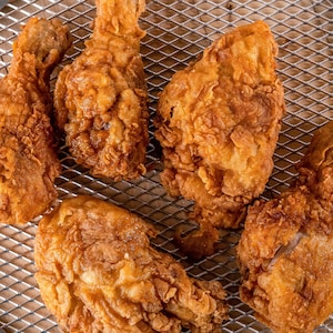 Peut inclure: Vue aérienne de cinq morceaux de poulet frit brun doré sur une grille de refroidissement en métal. Le poulet a un extérieur croustillant et texturé. L'arrière-plan est une surface en bois de couleur claire.