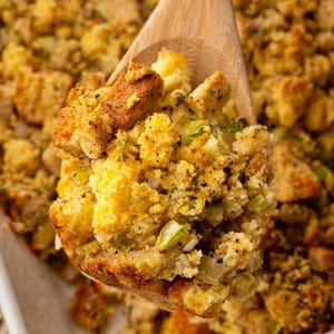 Puede incluir: Primer plano de una cuchara de madera que sostiene una porción de relleno. El relleno es una mezcla de cubos de pan dorados, hierbas y verduras. Condimentado con pimienta negra y especias, presenta una comida sabrosa y visualmente atractiva.