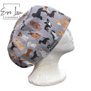May include: A light gray scrub cap featuring a repeating pattern of various dog breeds in shades of brown, black, white, and tan. The fabric cap includes a small gray tag on the side with the text Scribes and a logo in the upper left corner.