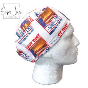 May include: A white scrub cap featuring a repeating graphic print of hot dogs with the text I GOT THAT, DOG IN ME, and 1.50. The fabric pattern includes red, blue, and yellow colors. A small grey rectangular tag with the word SCRIBES is sewn on the side.