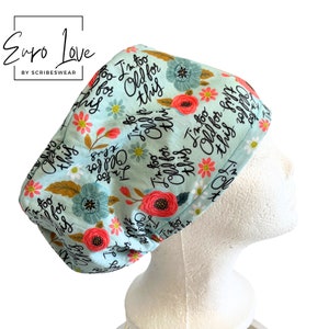 May include: A light blue scrub cap featuring a repeating floral pattern in shades of red, teal, and yellow, interspersed with the black cursive text "I'm too old for this." The fabric has a soft texture and is worn on a white mannequin head.
