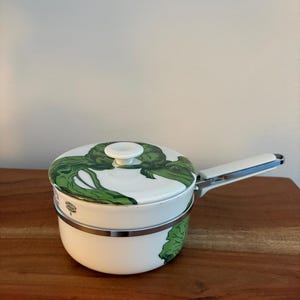 May include: White ceramic saucepan with a lid featuring a green artichoke design. The saucepan has a silver rim and a white handle with a blue accent. The handle is folded down. The saucepan is on a wooden surface.