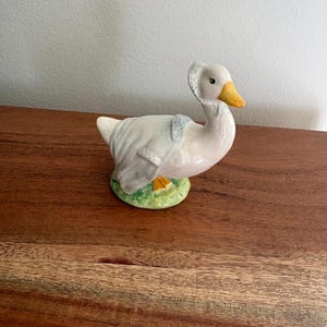 May include: A ceramic goose figurine with a white body, yellow beak, and a bonnet. The goose is wearing a light blue shawl and is standing on a small, round base of green grass. The figurine is a decorative item.