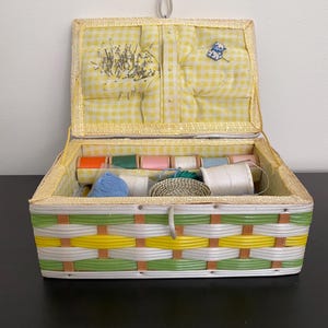 May include: A vintage sewing box with a yellow and white checkered fabric lining. The box is made of woven plastic with green, yellow, and white stripes. The box contains spools of thread, a ball of yarn, and a pincushion with pins.
