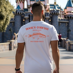 May include: White t-shirt with an orange print of a fish and the text "HOOK'S CODFISH RESTAURANT EST. 1953 PAN-FRIED DAILY".