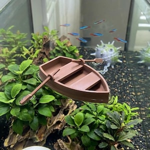 以下が含まれることがあります： 水槽用装飾品として設計された、2本のオールが入った小さな茶色のプラスチック製手漕ぎボート。ボートはベンチシートを備えたシンプルな船体の形状をしており、緑の水草と暗い色の砂利の間に配置されています。