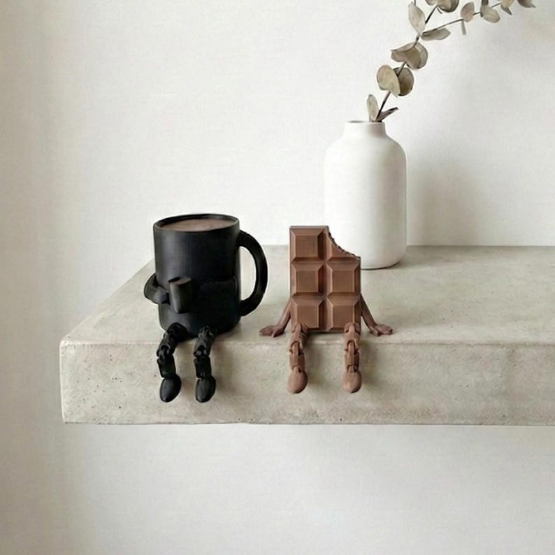 Pu&ograve; includere: Una tazza da caff&egrave; nera e una barretta di cioccolato, entrambe con gambe e braccia attaccate, sono posizionate fianco a fianco su uno scaffale. La tazza contiene un liquido scuro e la barretta di cioccolato ha un morso. Un vaso bianco con della vegetazione &egrave; sullo sfondo.