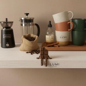 May include: A coffee-themed still life featuring a black coffee grinder, a French press, a burlap sack of coffee beans, and a stack of mugs. A bottle of vanilla extract and a chocolate bar figure are also present. The Lumi Store logo is visible.