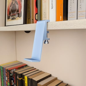 May include: A light blue metal shelf clamp, designed to attach to a shelf. The clamp has a flat base and a vertical support with a screw mechanism for securing it to the shelf. Books are visible in the background.