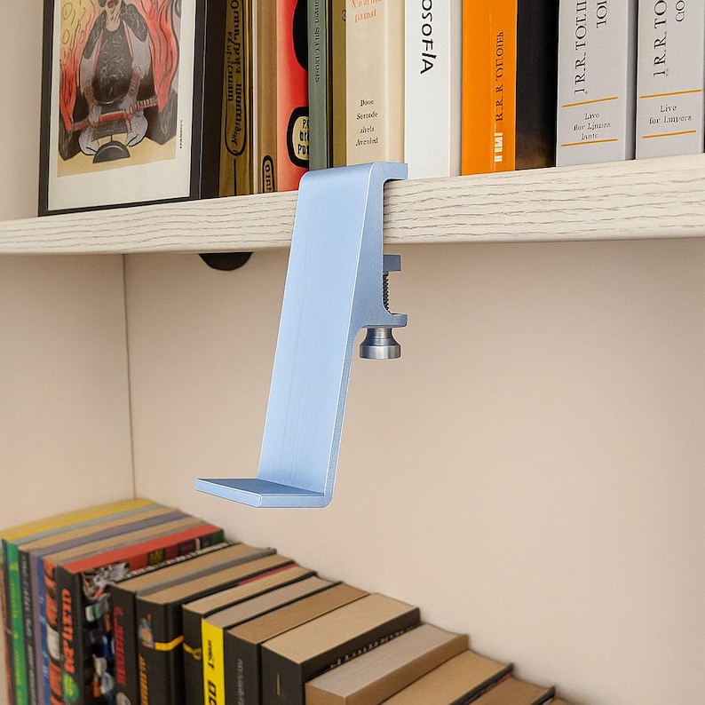 May include: A light blue metal shelf clamp, designed to attach to a shelf. The clamp has a flat base and a vertical support with a screw mechanism for securing it to the shelf. Books are visible in the background.